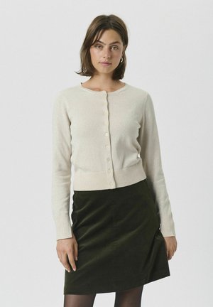 Hellbeige Cardigan mit Rundhalsausschnitt, vorderen Knöpfen und langen Ärmeln. Über einem olivgrünen Rock getragen, der aus einem strukturierten Stoff besteht.
