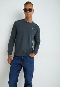 Camiseta de algodón de manga larga en gris oscuro con un pequeño logotipo blanco, combinada con jeans de mezclilla azul. Tejido texturizado con un ajuste relajado.