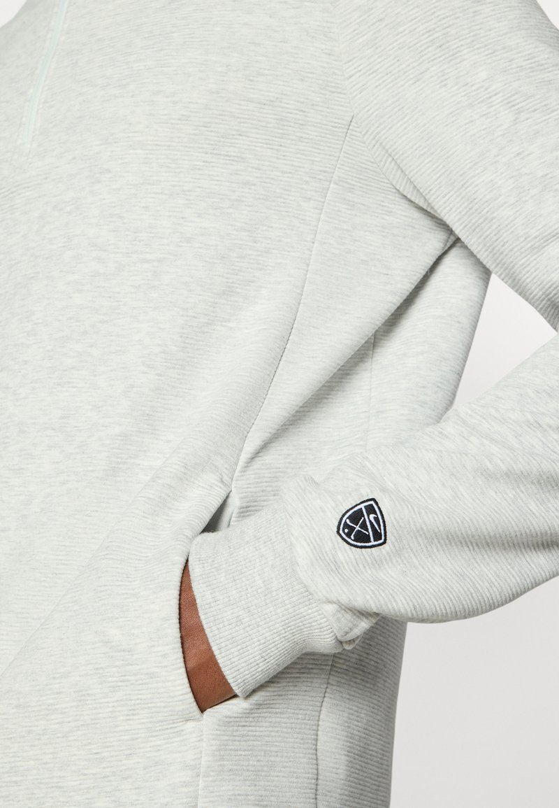 Hellgrauer Pullover aus strukturiertem Stoff, mit Viertel-Reißverschluss, gerippten Bündchen und einem kleinen schwarzen Emblem am Ärmel. Seitentaschen sichtbar.