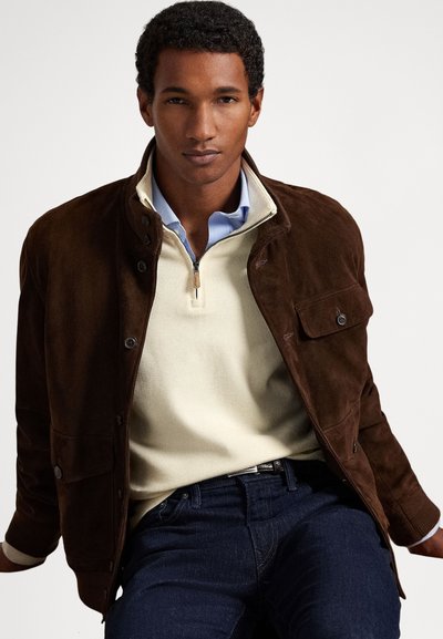 Muž v hnědém semišovém kabátu přes béžový svetr se zipem a modrou košilí s límečkem, sedící před jednobarevným sv�ětlejším pozadím.