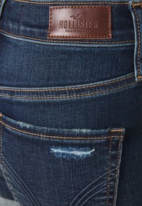 Džíny z tmavě modrého denimu s hnědou koženou značkou "Hollister California." Poškozený detail kapsy a kontrastní oranžové švy.