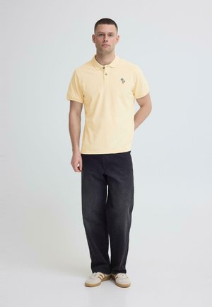 Homme portant un polo jaune clair avec un petit logo de palmier, un jean noir et des baskets blanches, debout devant un fond clair uni.