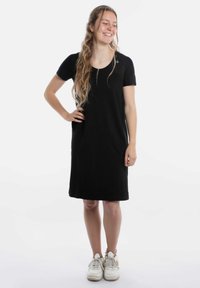 Zwarte jurk met korte mouwen en een ronde halslijn, voorzien van zijzakken, gemaakt van zacht materiaal; gedragen met witte sneakers. Heeft een minimalistisch ontwerp.