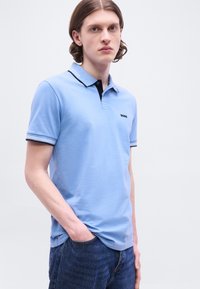 Νεαρός άνδρας με μαλλιά μέχρι τους ώμους φοράει ανοιχτό μπλε πόλο πουκάμισο με μαύρο περιθώριο και σκούρο μπλε τζιν, στέκεται με το ένα χέρι στην τσέπη.