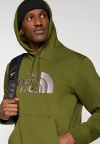 Φούτερ με κουκούλα σε ελιά αποχρώσεις, κατασκευασμένο από βαμβακερό μείγμα, με μεγάλο κεντημένο λογότυπο στο στήθος, μπροστινή τσέπη και ρυθμιζόμενη κουκούλα με κορδόνι.