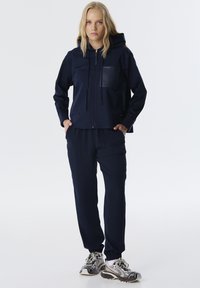 Chaqueta con capucha en color azul marino, con un bolsillo frontal, combinada con pantalones holgados a juego. El material parece suave y ligeramente brillante. Presenta puños elásticos.