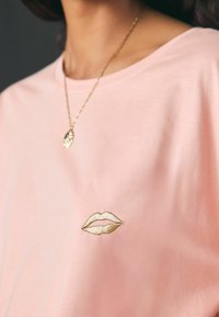 Šviesiai rožinė medvilnės marškinėliai su auksine siuvinėta lūpų dizainu ant krūtinės. Minkšta tekstūra, laisvas kirpimas ir minimalistinis detalizavimas.