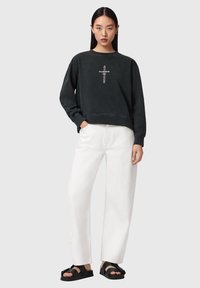 Kvinde med langt sort hår iført sort sweatshirt med lodret AllSaints-logo, hvide vidde bukser og sorte sandaler med åben tå, hænderne i lommerne.