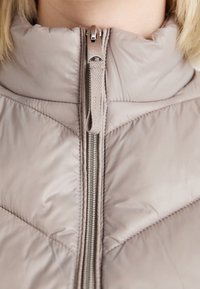 Gilet rembourré en taupe clair avec un col haut. Doté d'une fermeture éclair argentée et d'un motif de coutures en chevrons sur le devant. Texture lisse et brillante.