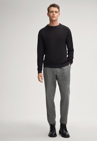 Czarny sweter o fakturze, szare spodnie w kratę i czarne sznurowane botki. Model stoi z rękami w kieszeniach na neutralnym tle.