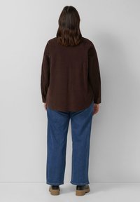 Bruine blouse met lange mouwen, afgeronde zoom; gecombineerd met losse blauwe jeans en zwarte boots met dikke zolen.