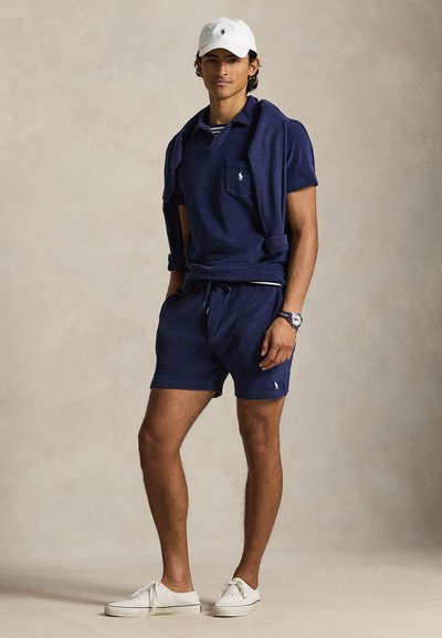 Μπλουζάκι πόλο σε ναυτικό μπλε με τσέπη, συνδυασμένο με σορτς σε ίδια απόχρωση. Φοριέται με λευκό καπέλο και αθλητικά παπούτσια, παρουσιάζοντας ένα casual καλοκαιρινό ύφος.