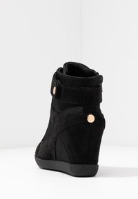 Černé wedge tenisky s botou, vyrobené z tkaniny a semiše, s páskem kolem kotníku, zlatým knoflíkovým akcentem a gumovou podrážkou na bílém pozadí.