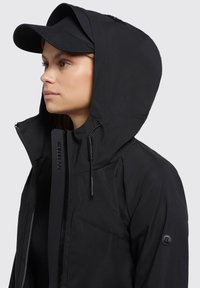 Giacca nera da esterno con zip frontale, cappuccio regolabile e tasche. Tessuto morbido con dettagli hardware sottili. Berretto indossato in cima alla testa.
