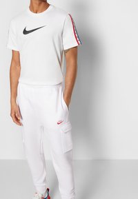 Nike vit t-shirt med svart logotyp, med röda och blå ränder på ärmarna; kombinerad med vita cargo-byxor med röd logotyp och fickor.