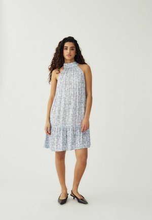 Junge Frau mit langen lockigen Haaren, die ein ärmelloses, hochgeschlossenes, helles Blumenkleid und schwarze spitze Schuhe mit niedrigem Absatz trägt und vor einem weißen Hintergrund steht.