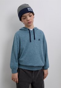 Hanorac albastru tricotat cu buzunar frontal și șnururi bleumarin, asortat cu o căciulă gri în dungi cu o etichetă logo. Pantaloni din corduroi gri texturat.