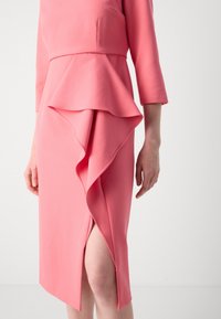 Robe ajustée rose avec un détail de volants superposés à la taille, des manches longues et une fente haute sur un côté. Texture de tissu douce.