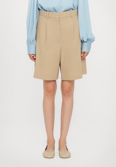 Beige maßgeschneiderte Shorts mit Falten, aus leichtem Material gefertigt. Getragen mit einem hellblauen Langarmshirt und beigen Ballerinas.