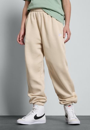 Pantalons de survêtement beiges avec taille élastique et poignets, texture douce, arborant un petit logo blanc Nike sur la cuisse gauche. Associés à des chaussures montantes blanches.