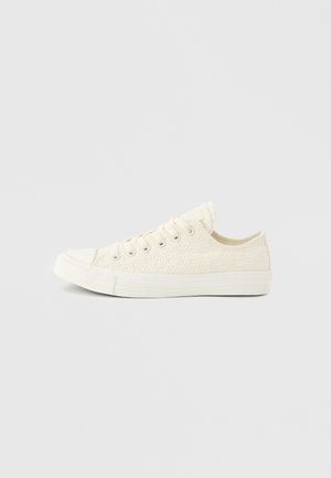 Άσπρο sneaker χαμηλού πέλματος με υφή δαντέλας, λευκό λάστιχο στη σόλα και λευκά κορδόνια σε ανοιχτό γκρι φόντο.