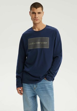 Młody mężczyzna w granatowej koszuli z długim rękawem z graficznym prostokątnym nadrukiem i jasnoniebieskich dżinsach, stojący z jedną ręką w kieszeni.