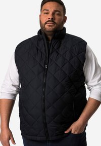 Černá prošívaná vesta s vysokým límcem, zapínáním na zip a dvěma bočními kapsami. Nositelná přes bílou košili s dlouhým rukávem, která má texturovaný povrch.