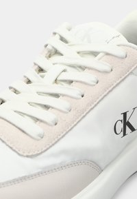 Primo piano di una sneaker bianca e beige con lacci bianchi e logo "cK" sul lato.