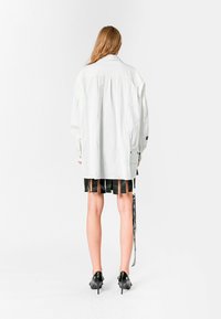 Camisa blanca de gran tamaño con un sutil detalle texturizado, falda negra con plisados y zapatos de tacón negros. Etiqueta visible en la falda.