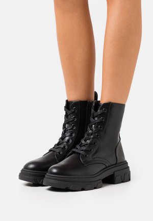 Jambes portant des bottines à lacets épaisses noires avec fermetures éclair sur le côté, vues de face et du côté extérieur sur fond blanc.