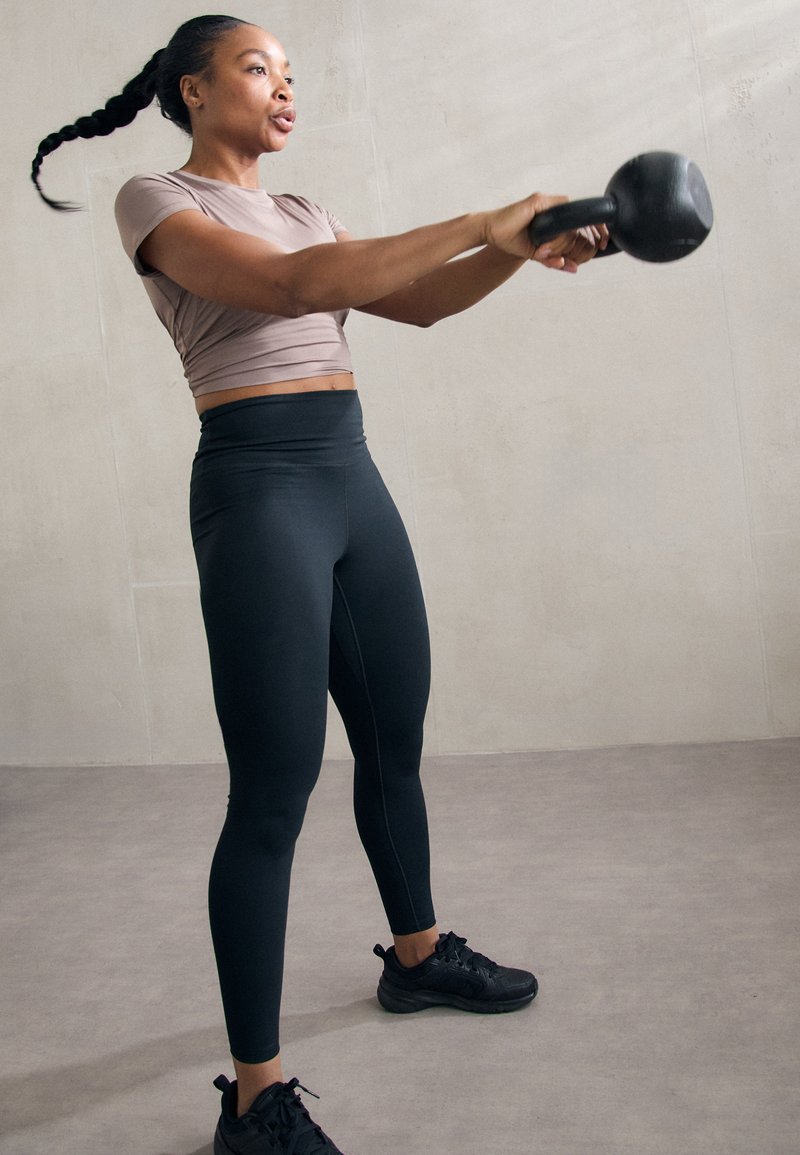 Zwarte high-waisted legging en een lichtbruine cropped top. Het model zwaait met een kettlebell, wat beweging en atletisch vermogen benadrukt.