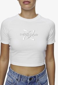 Hvit kortermet t-skjorte med sølvfarget "PEGADOR"-logo. Kombinert med høy midje, blå denimshorts. Glatt tekstur.