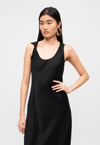 Μαύρο σατέν φόρεμα με στρογγυλεμένο ντεκολτέ, με μια λεπτομέρεια λουριού που στρίβει σε έναν ώμο. Λείο ύφασμα και ρέων σχέδιο.