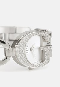 Montre-bracelet argentée avec un cadran blanc, ornée de cristaux et d'un logo en évidence. Présente un design de bracelet à maillons.