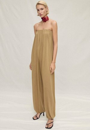 Tuta beige senza spalline con tessuto fluente, design a gamba ampia e vita strizzata. Completata da un choker in tessuto rosso e sandali neri.