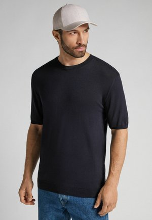 Marineblå kortærmet sweater med rund halsudskæring, ribbet kant og lysegrå baseballcap. Blød, glat tekstur, båret med blå jeans.