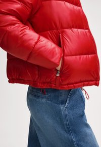 Czerwona kurtka ocieplana z błyszczącym wykończeniem, z boczną kieszenią na zamek błyskawiczny, elastycznym wykończeniem u dołu i regulowanymi sznurkami po bokach. W połączeniu z niebieskimi dżinsami.