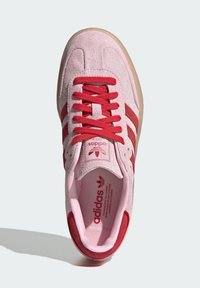 Roze suede sneaker met rode veters en drie rode strepen. Beschikt over een gewatteerde kraag en een getextureerde gumrubberen zool. Adidas branding zichtbaar.