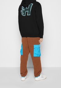 Felpa nera con un grande logo "H" in teal, abbinata a pantaloni in pile marroni con tasche cargo blu. Le sneakers bianche completano l'outfit.