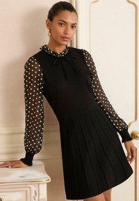 Black dress with a fitted bodice and pleated skirt. Sheer, polka-dotted long sleeves with ruffled cuffs and a bow tie at the neckline.