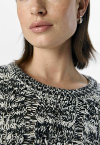Sweter dziany z czarno-białym wzorem, okrągłym dekoltem, teksturowanym materiałem i widocznymi detalami szycia. Prezentowane są kolczyki na ucho.