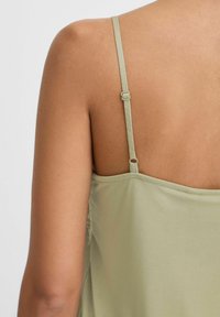 Camisole verde chiaro con sottili spalline regolabili, caratterizzato da una superficie liscia e una caduta morbida. Include un bordo attorno al collo.