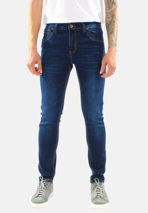 Uomo che indossa jeans slim fit blu scuro, t-shirt bianca, sneakers grigie, e mostra un tatuaggio sul avambraccio sinistro.