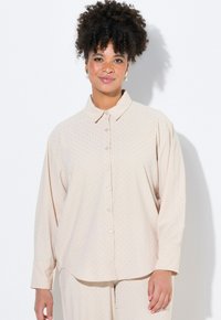 Blusa de color beige claro con un cierre de botones en la parte delantera, mangas largas y un sutil patrón de lunares. Presenta un cuello clásico y una textura de tela suave.