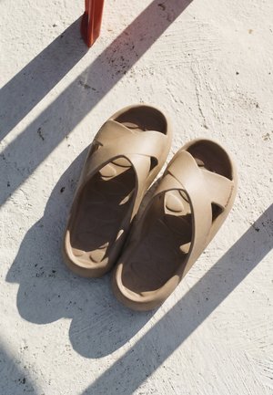 Paar hellbraune Slip-On-Sandalen mit gekreuzten Riemen, die auf einer strukturierten weißen Oberfläche mit nahegelegenen Schatten liegen.
