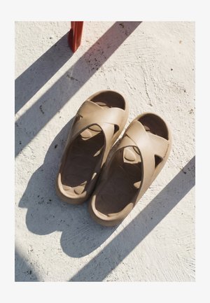 Paar hellbraune Slip-On-Sandalen mit gekreuzten Riemen, die auf einer strukturierten weißen Oberfläche mit nahegelegenen Schatten liegen.