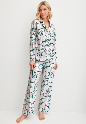 Vrouw staat op blote voeten en draagt een pyjamaset met een langarmige, knoopsluitende top en wijde broek met groot wit-groen bloemenpatroon.