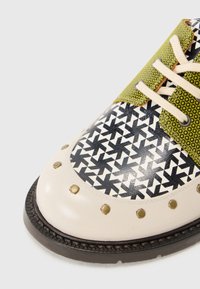 Multicolored shoe featuring a black and white geometric pattern, green accents, beige leather, gold studs, and beige laces.