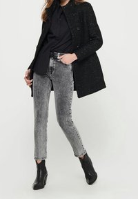 Schwarzer, strukturierter Mantel mit Knöpfen, kombiniert mit grauen, hoch taillierten Skinny-Jeans. Schwarze Stiefeletten runden den Look ab.