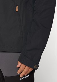 Zwarte waterdichte jas met een ritssluiting zak, tan leren accenten en een knoopsluiting aan de mouw. Glad materiaal met een gestructureerd ontwerp.